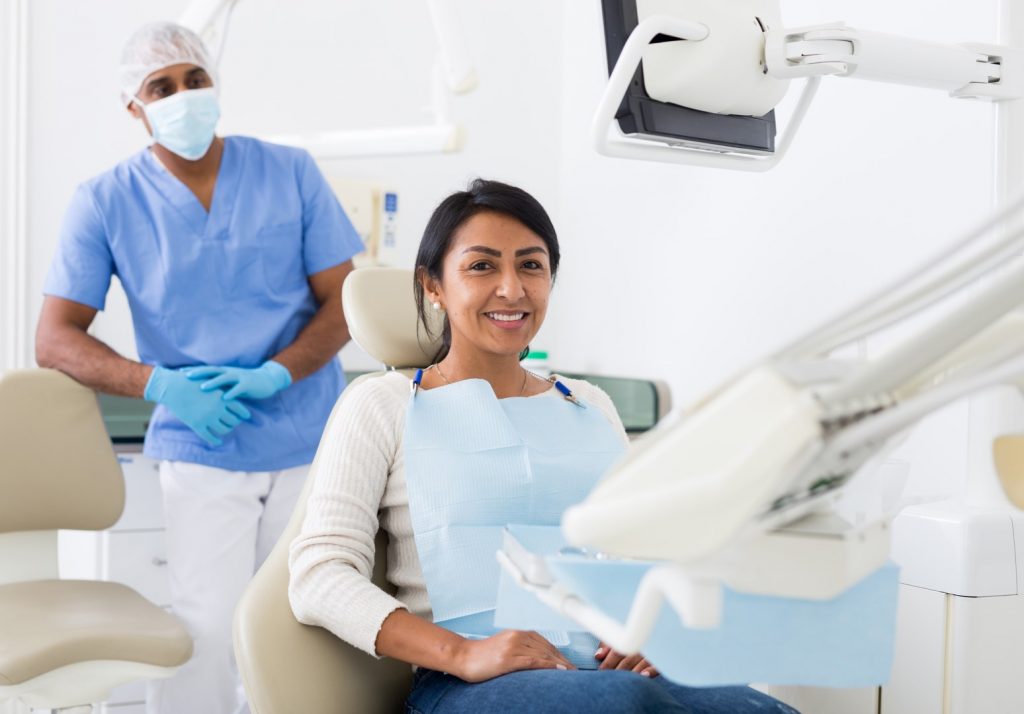 A woman in a dental chair smiles after a masked and gloved dentist behind her has found no signs of common dental problems.