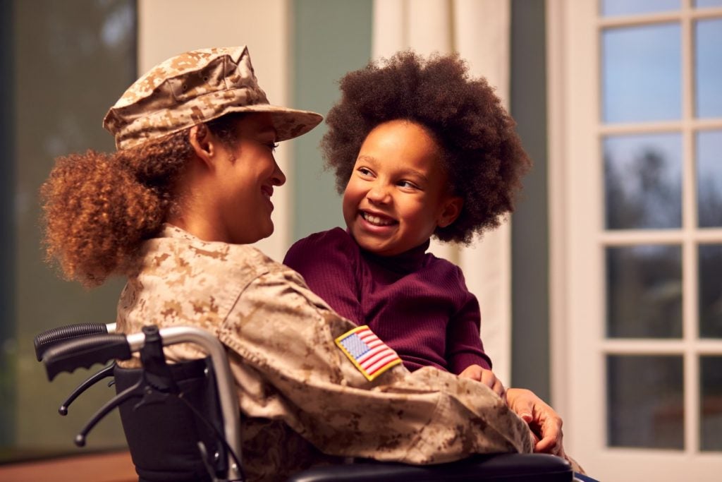 A veteran wearing a U.S. Army camo cap and jacket smiles at her daughter, who sits on her lap.