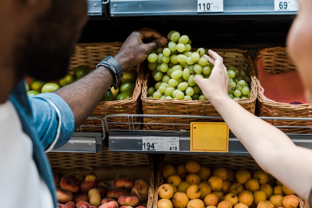 A man and woman pick a bunch of grapes from a grocery store produce section because you can eat grapes with braces.