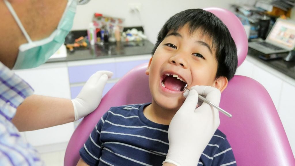 An elementary school-age boy reclines in a dental chair as an affordable children’s dentist in Philadelphia examines his teeth.