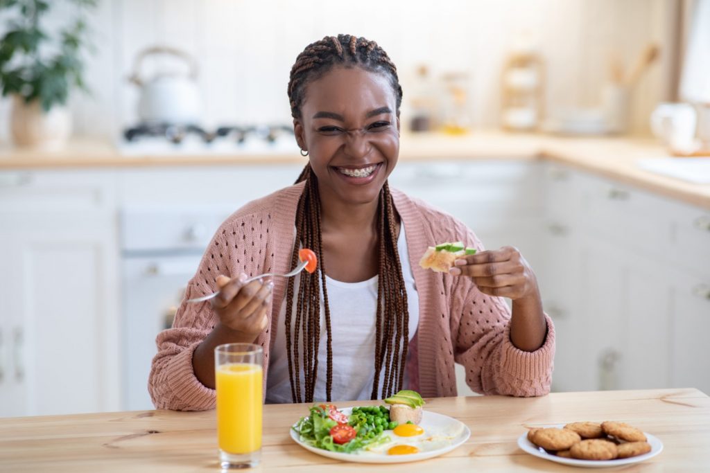 A woman with braces sits in her kitchen smiling as she eats a meal with fruits and vegetables, examples of what you can eat with braces.