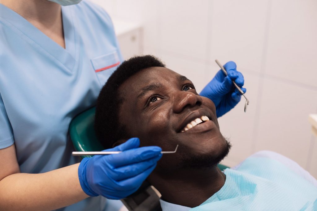 A man reclining in a dental chair smiles as a dentist prepares to examine his teeth using a dental pick and mirror.