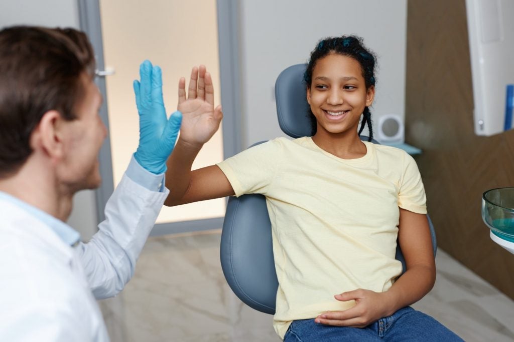 A teenage girl sits in a dental chair and gives a gloved dentist a high five after receiving affordable pediatric dental care.