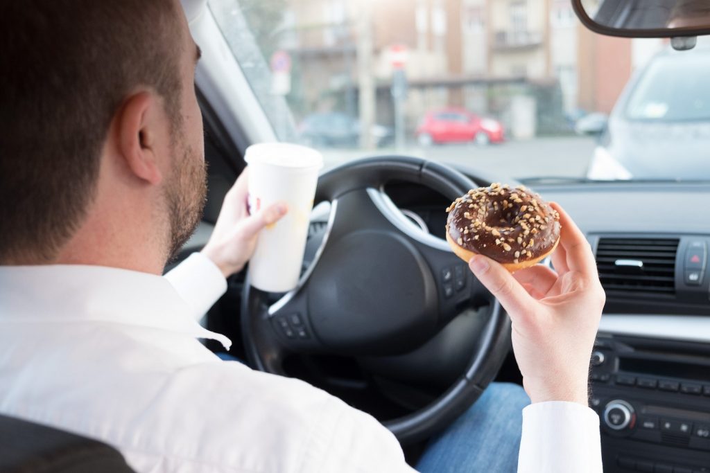 A man drinks sugary coffee and eats a chocolate-covered doughnut in his car—drink and food that’s not good for diet and oral health.