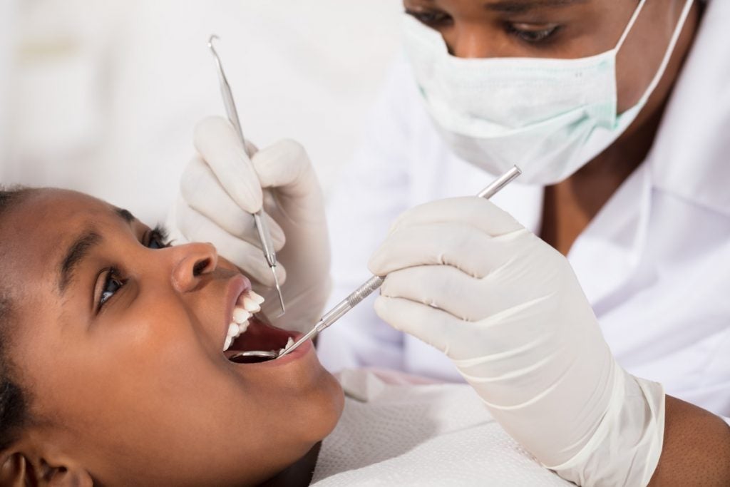 An elementary school-age girl reclines in a dental chair as an affordable children’s dentist examines her teeth with a mirror and pick.
