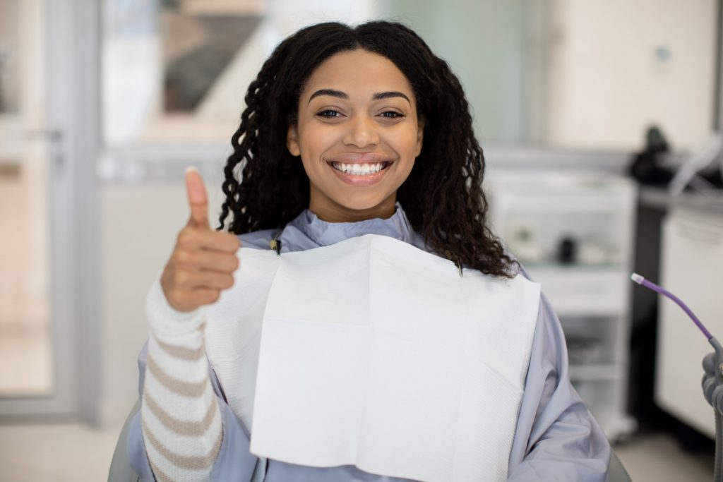 A happy young woman gives a thumbs-up and smiles from a dentist's office exam chair after her prosthodontist appointment. 