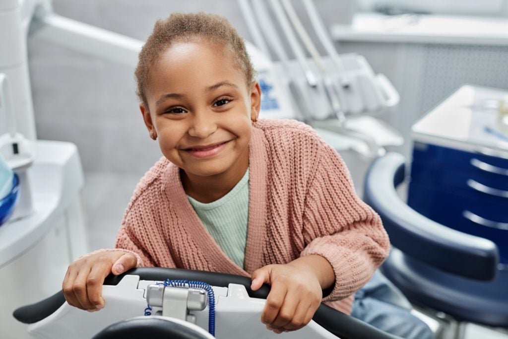 An elementary school-aged girl leans over the side of a dental chair and smiles after a pediatric dental sealants treatment.