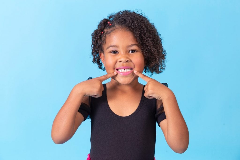An elementary school-aged girl points to her mouth and smiles after a pediatric dental sealants treatment.