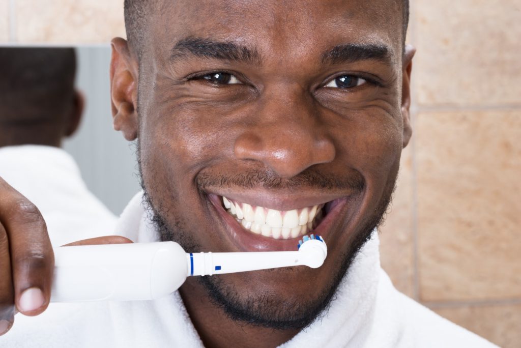 A man smiles while brushing his teeth with an electric toothbrush, an affordable dental health