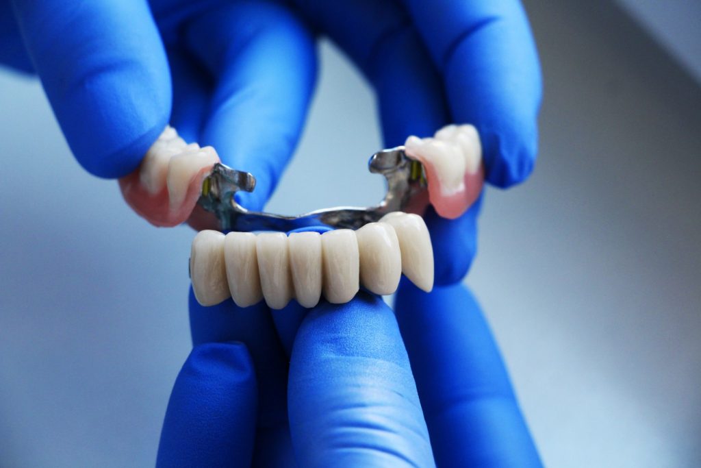 An affordable prosthodontist holds an affordable dental bridge in gloved hands before placing it in a patient’s mouth.