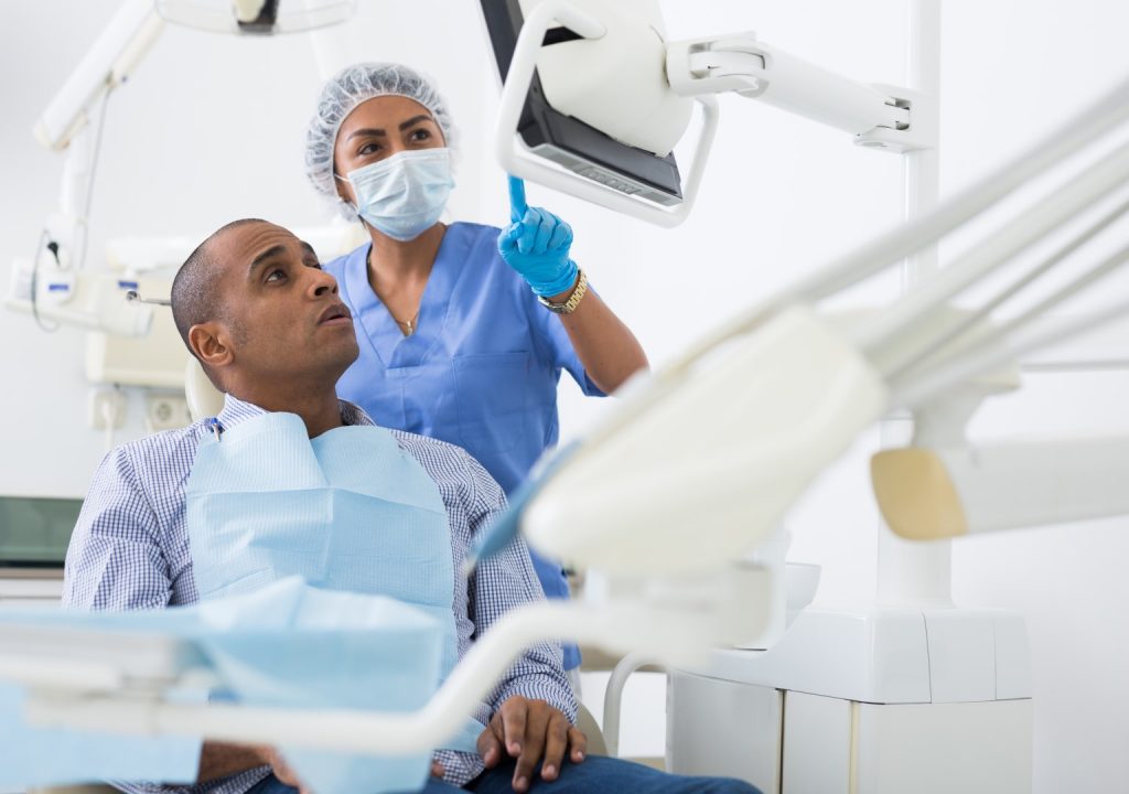 A female dentist wearing a mask points at a monitor to explain salivary gland disorders to a male patient. 