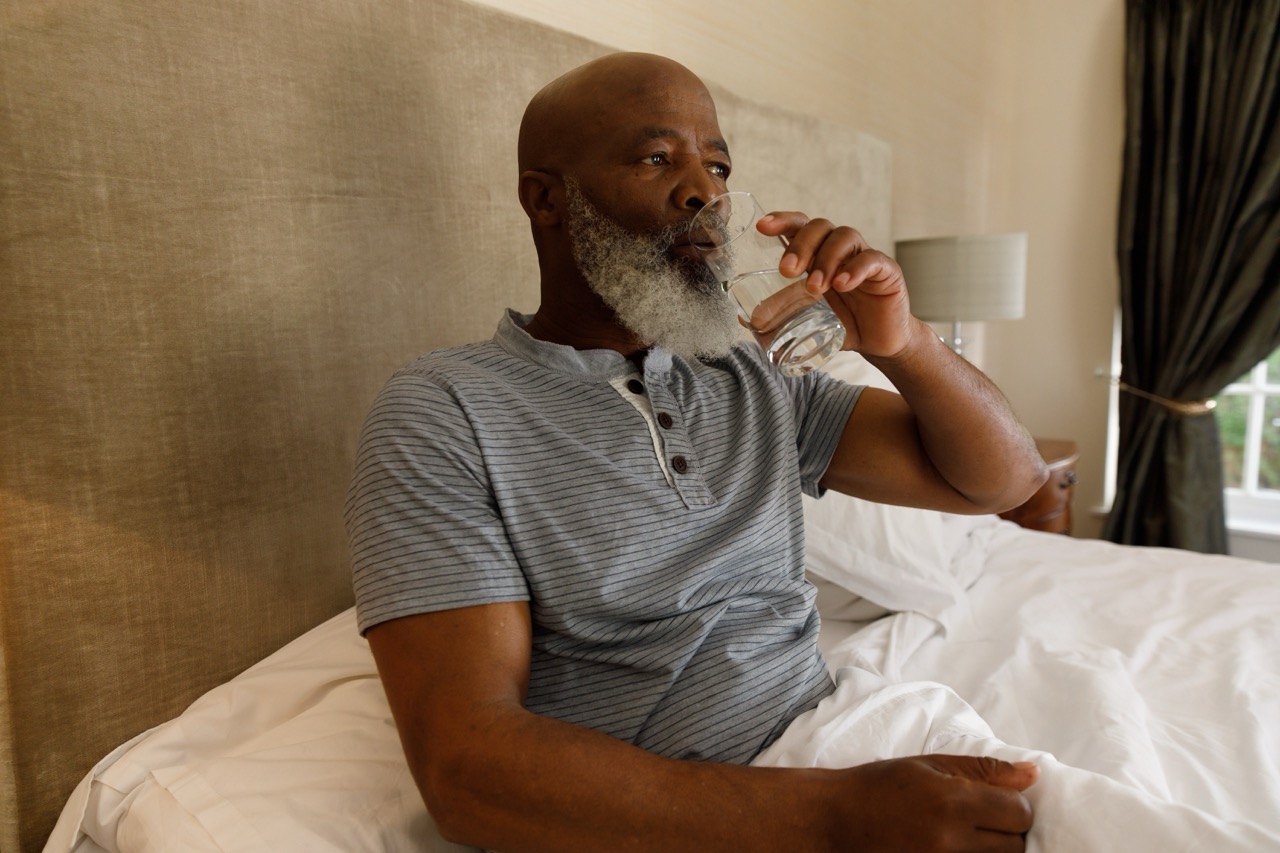 Man sits up in his bed to sip a glass of water as part of his home care for his dry mouth (xerostomia).