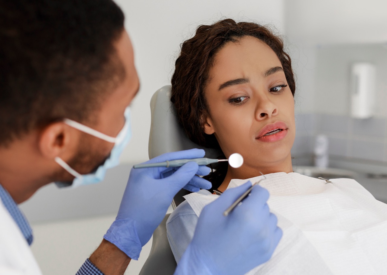 A scared young woman suffers from dental fear as she looks at the dental tools in the dentist’s hands.