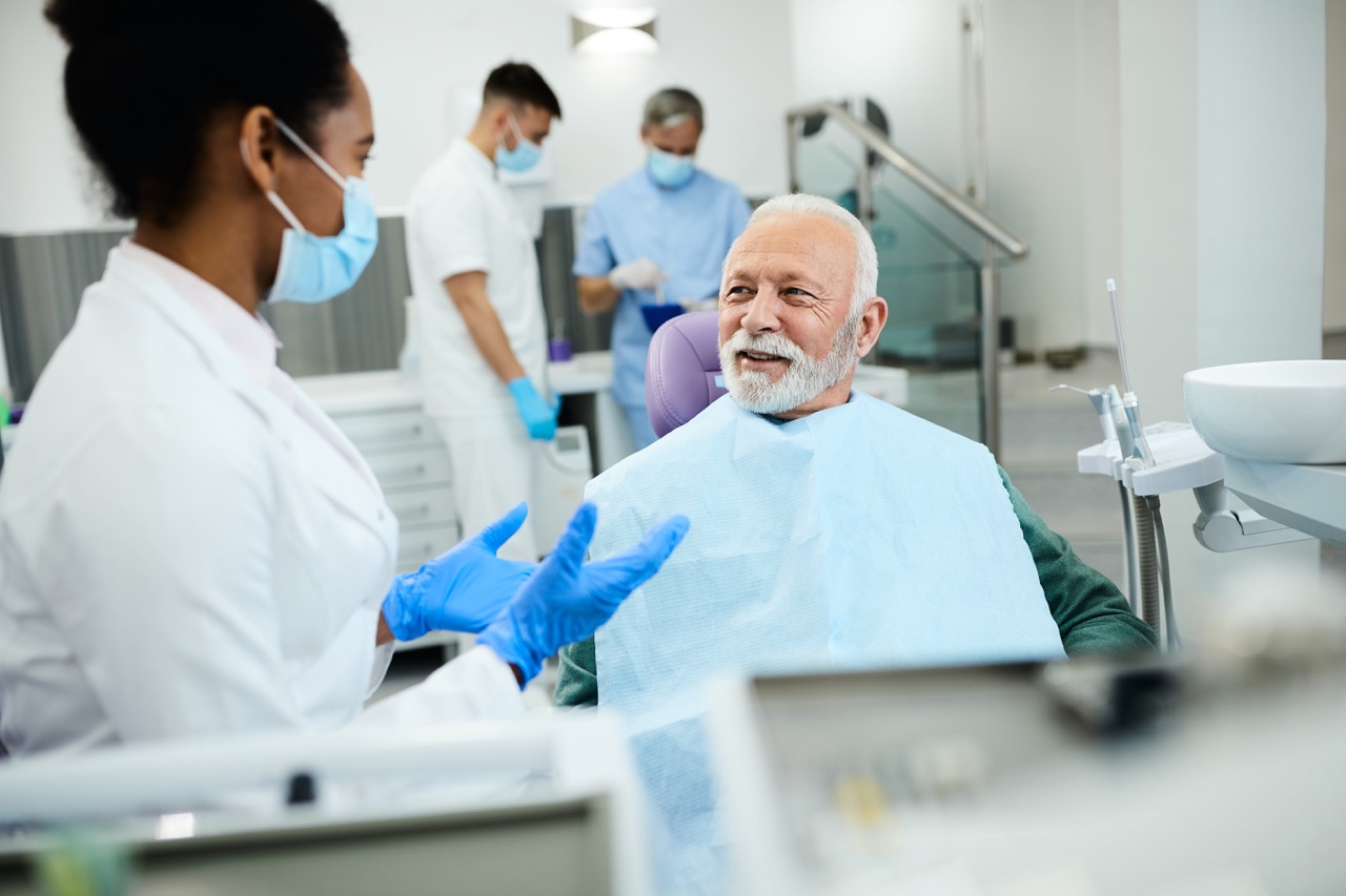 Black woman student dentist, wearing mask and gloves, talks with patient who sits in dental chair before treatment.