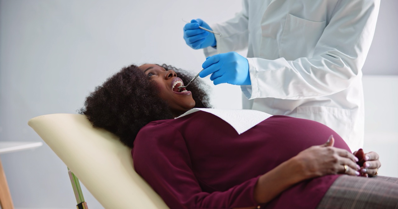 Pregnant woman reclines in dental chair as gloved dentist examines her teeth.