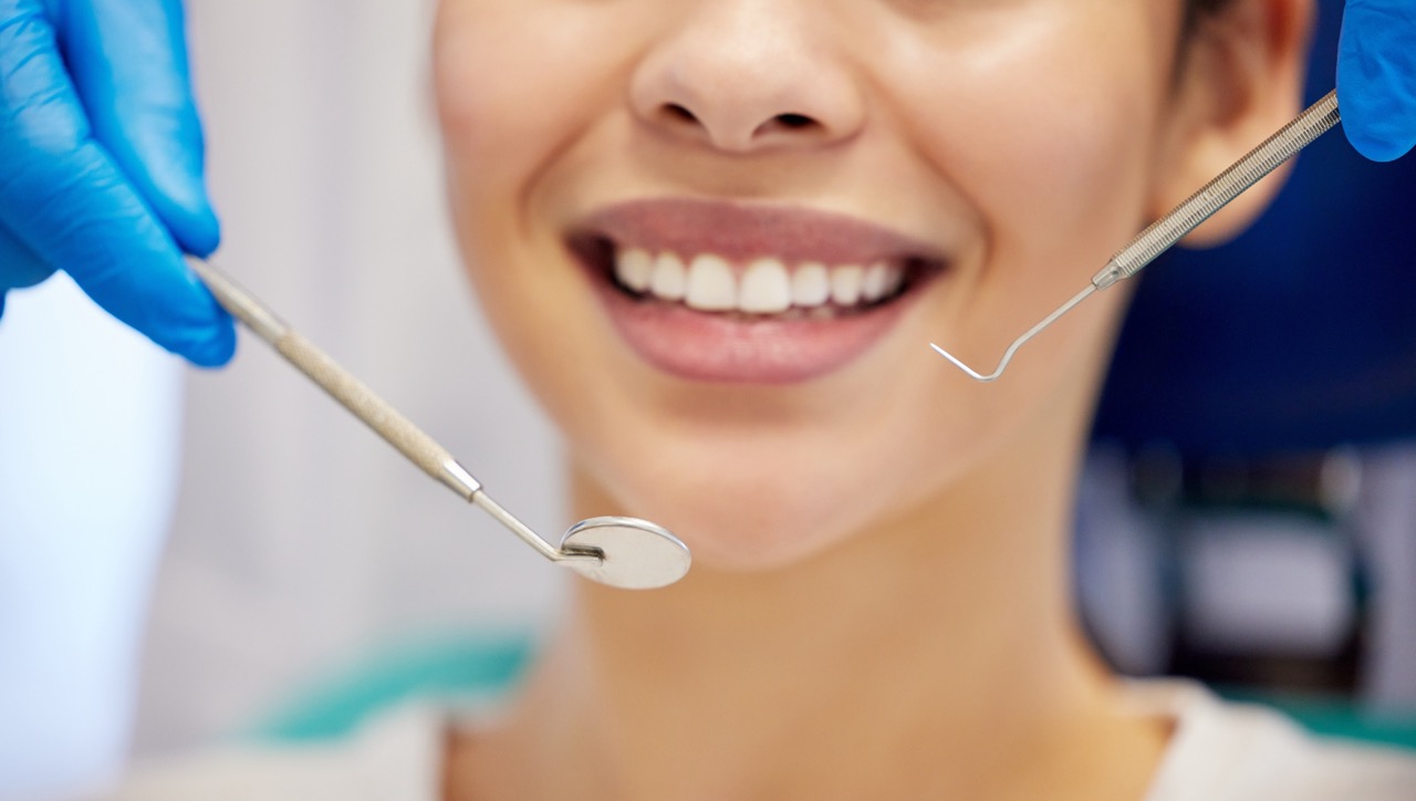 Student dentist’s gloved hands hold dental pick and mirror to mouth of woman in dental chair before routine examination.