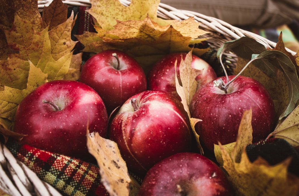 Six large red apples in a wicker basket, surrounded by autumn leaves, a teeth-friendly Halloween snack.