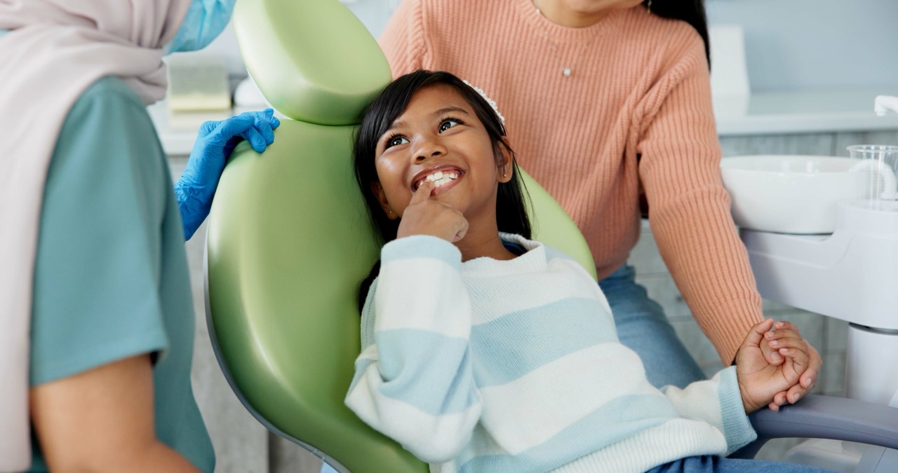 Image 3 Alt Text: A little girl sits in a dental chair and smiles at her dentist. 