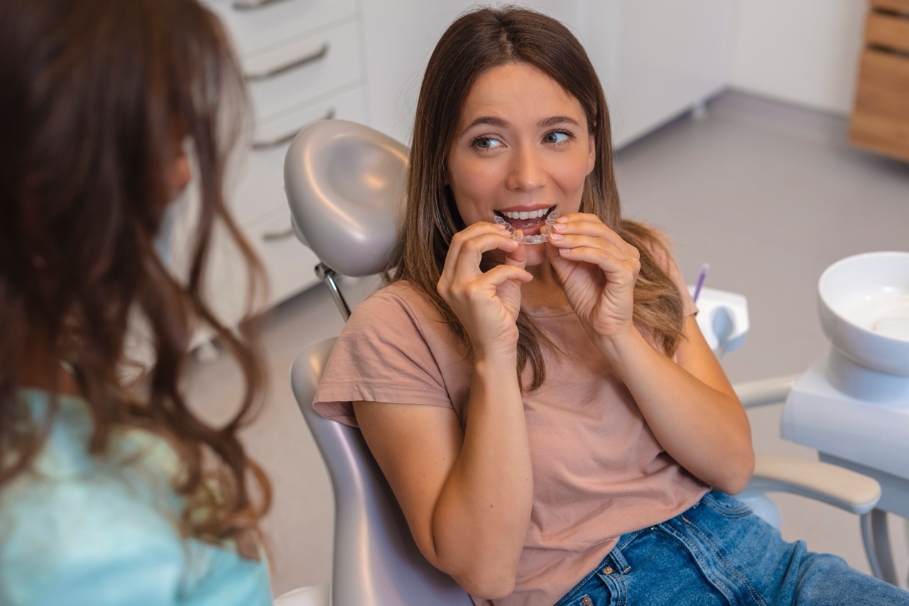 A young woman sits in the exam chair at the orthodontist’s office and holds a clear aligner to her teeth. 