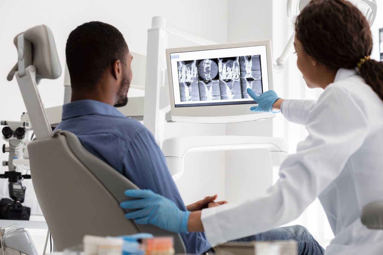 A man sitting in a dental exam chair looks at X-rays of his teeth with his dentist. 