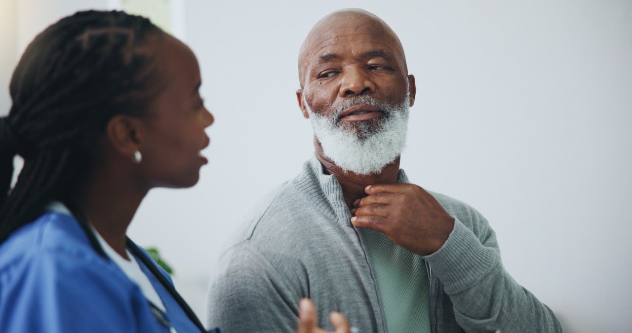 A dentist speaks to an older gentleman about oropharyngeal cancer systems as he holds his hand to his throat. 
