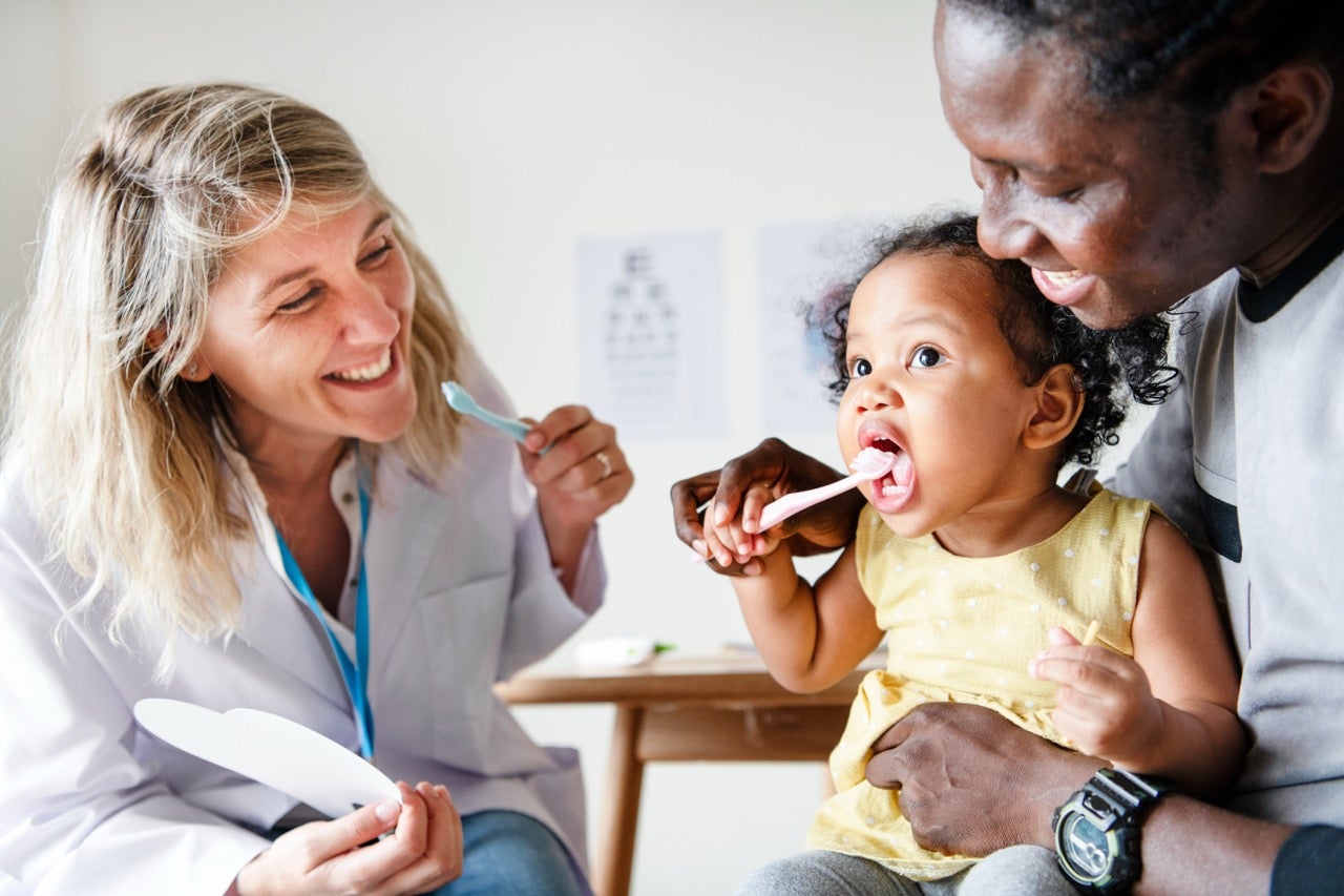 Female dentist helps teach a toddler how to brush her teeth as her dad holds her. 