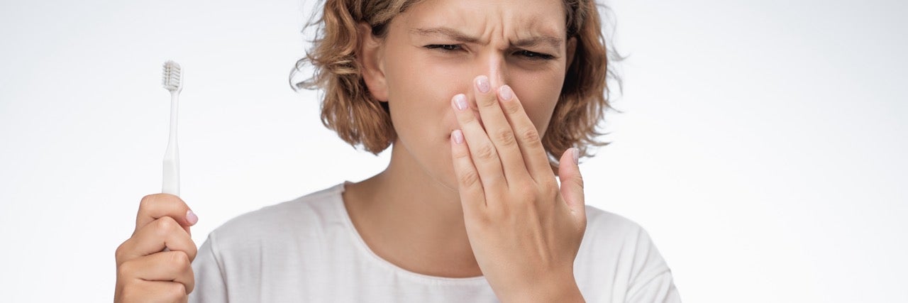 A young woman holds her hand up to her mouth to smell her breath while brushing her teeth. 