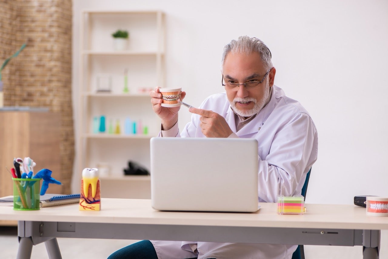 Dentist holds and points to model teeth with metal braces, communicating with his patient via laptop computer in teledentistry visit.