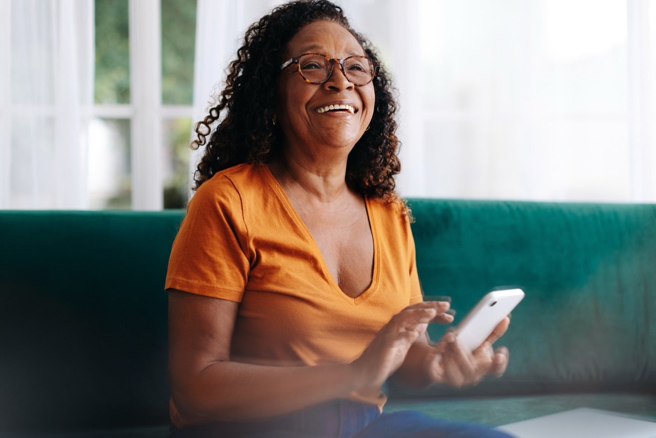 Woman sits on couch holding smartphone, smiling after a successful dental teledentistry consult.