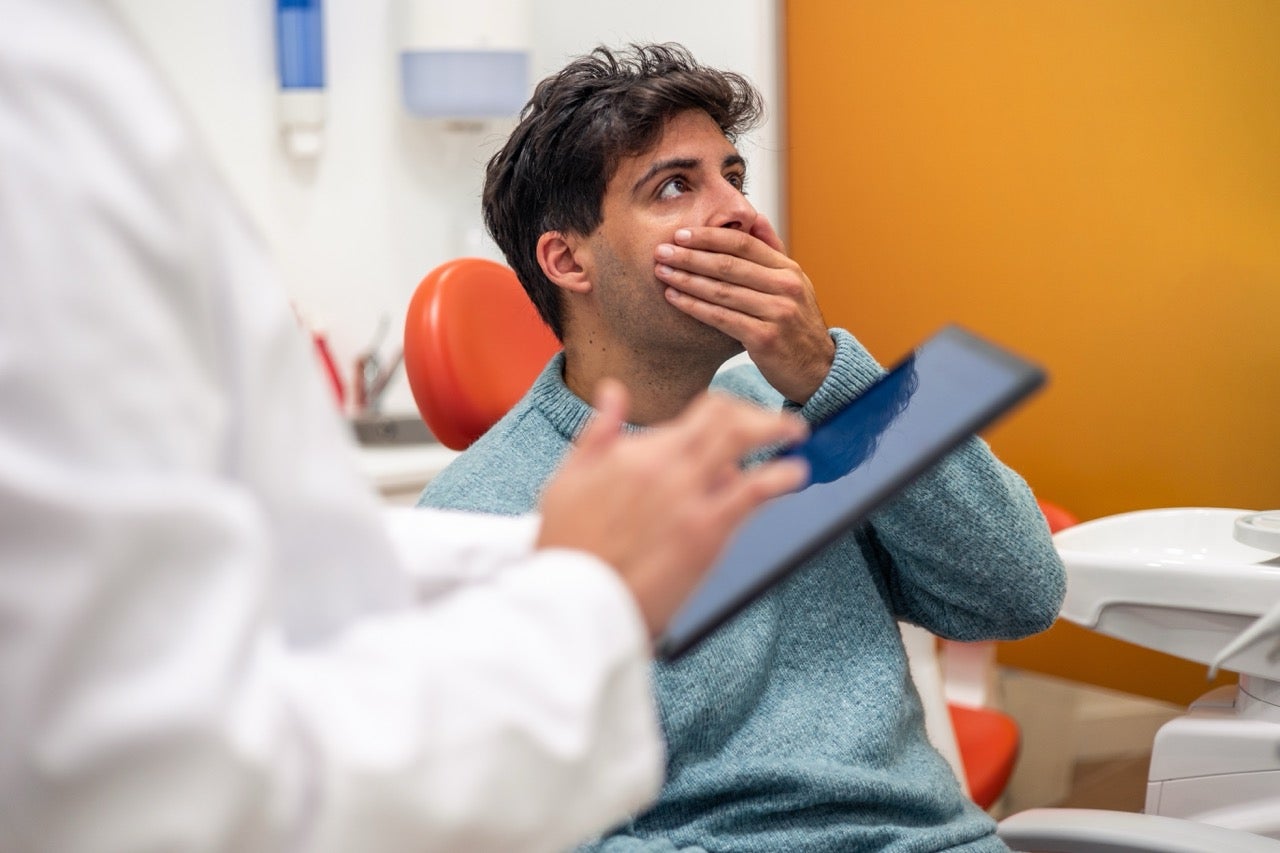 A young man is nervous and holds his hand to his mouth as his dentist shares information about his teeth.