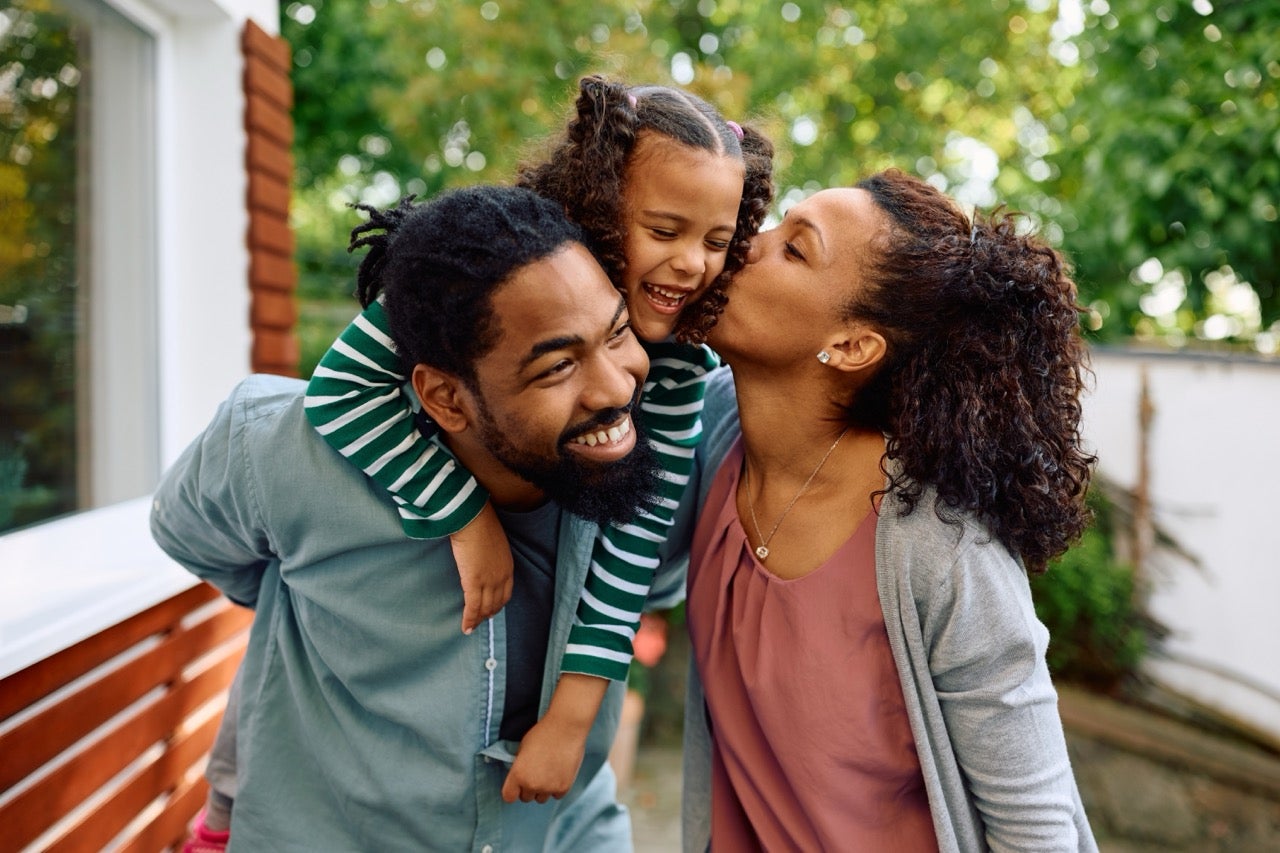 A young dad holds his daughter on his back, and her mom leans in to kiss her. 