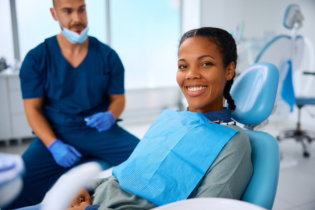 A young woman sits in the exam chair at the dentist’s office while the dentist looks on. 