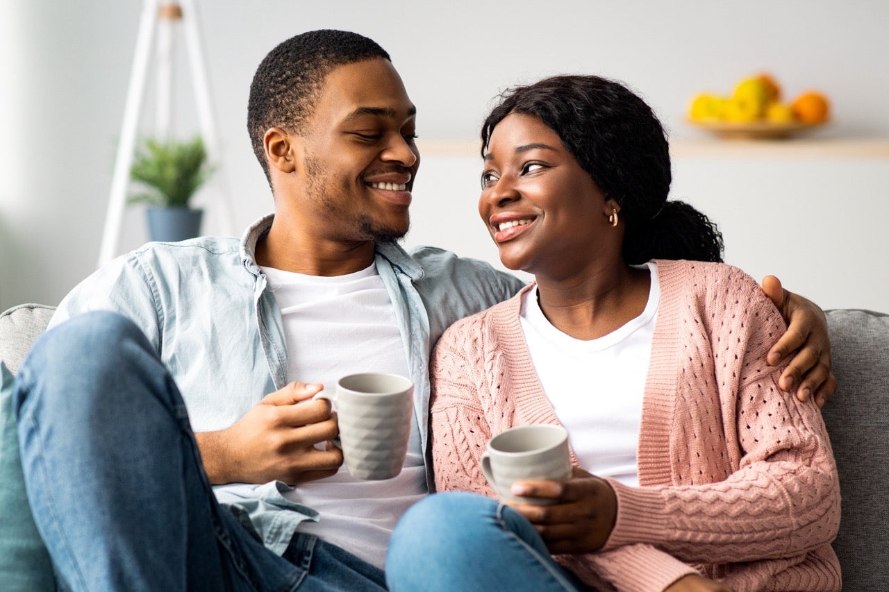 A young couple looks at each other while drinking coffee and sitting on the couch. 