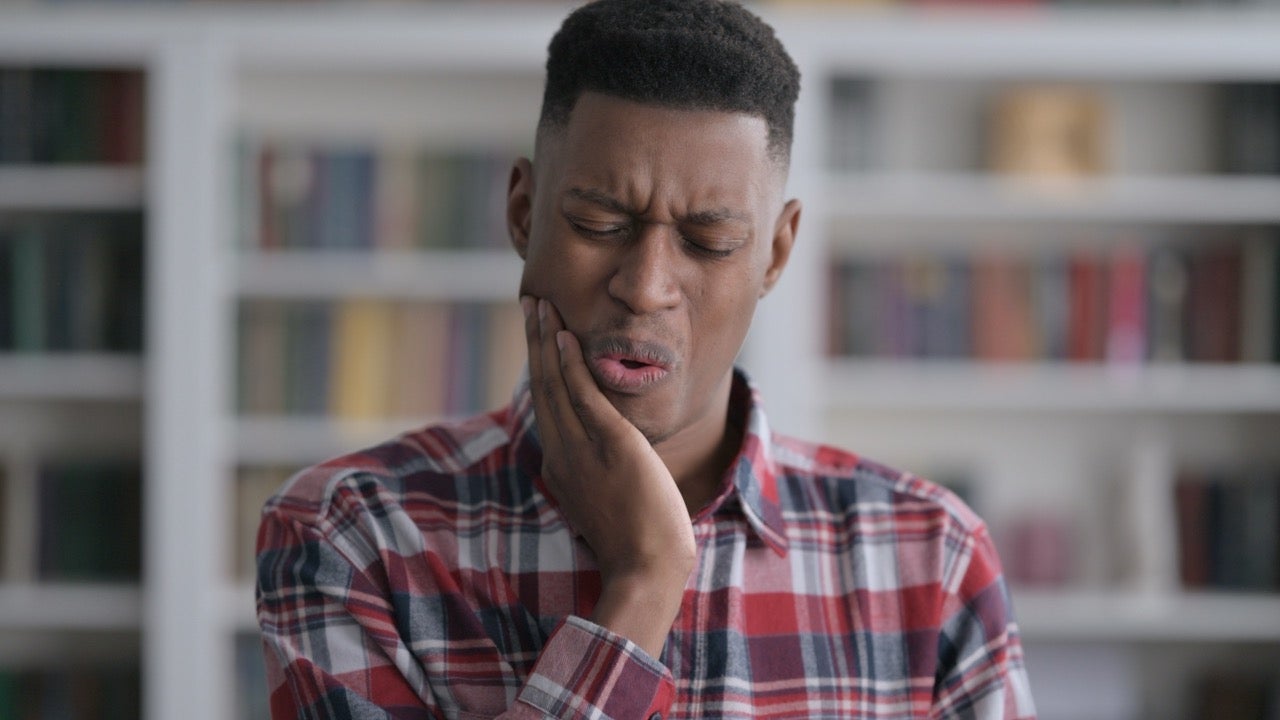 A young man wearing a plaid shirt has a toothache and holds his hand up to his mouth.