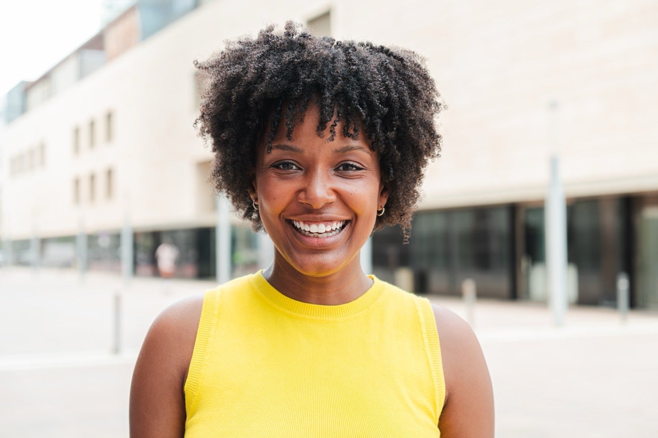 A smiling young woman in a bright yellow top standing in front of an office building. 