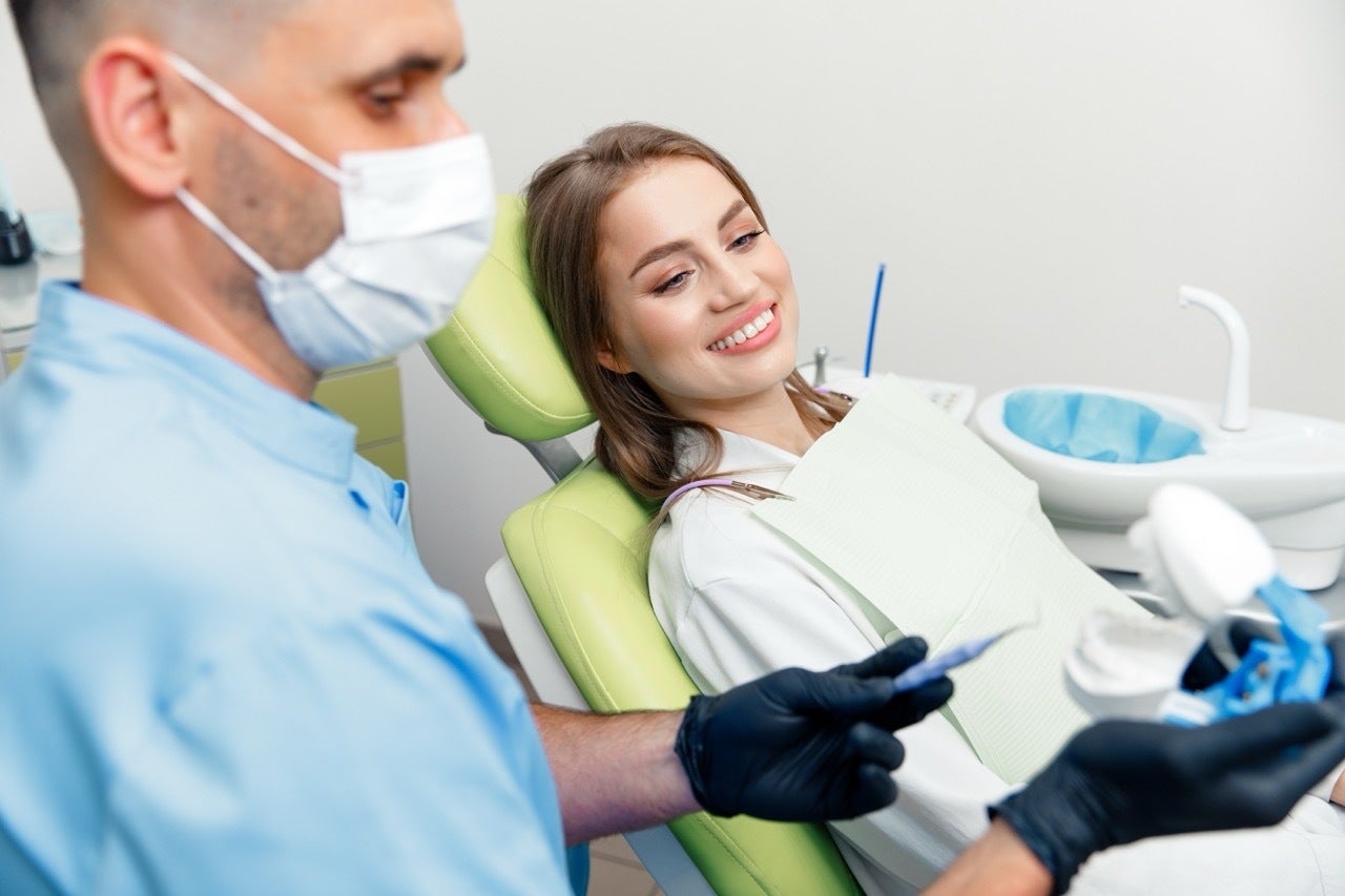 Masked dentist holds model of human teeth while explaining filling procedure to woman reclining in dental chair.