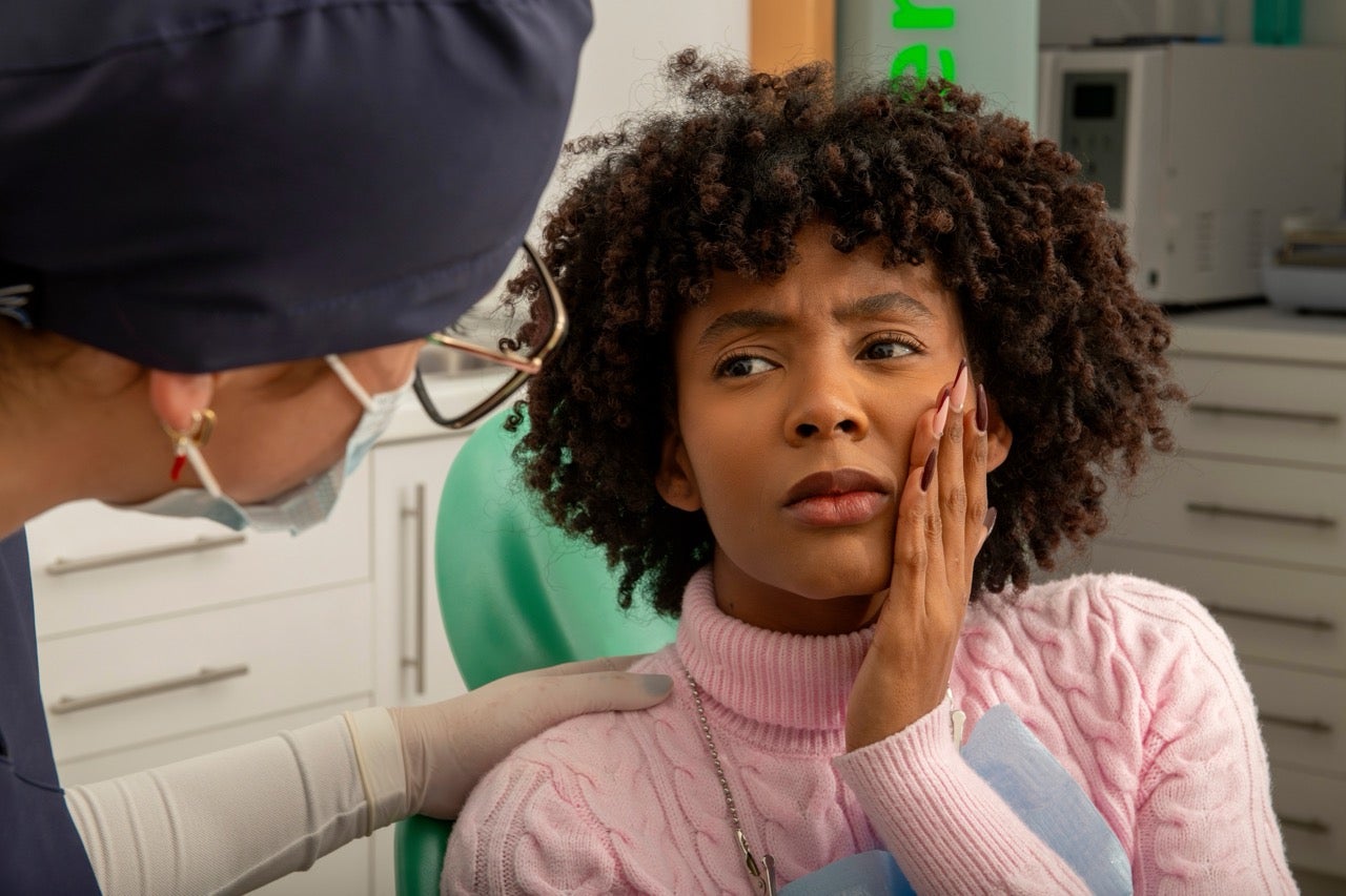 A young woman holds her hand to her face as she speaks to the dentist about gum disease. 
