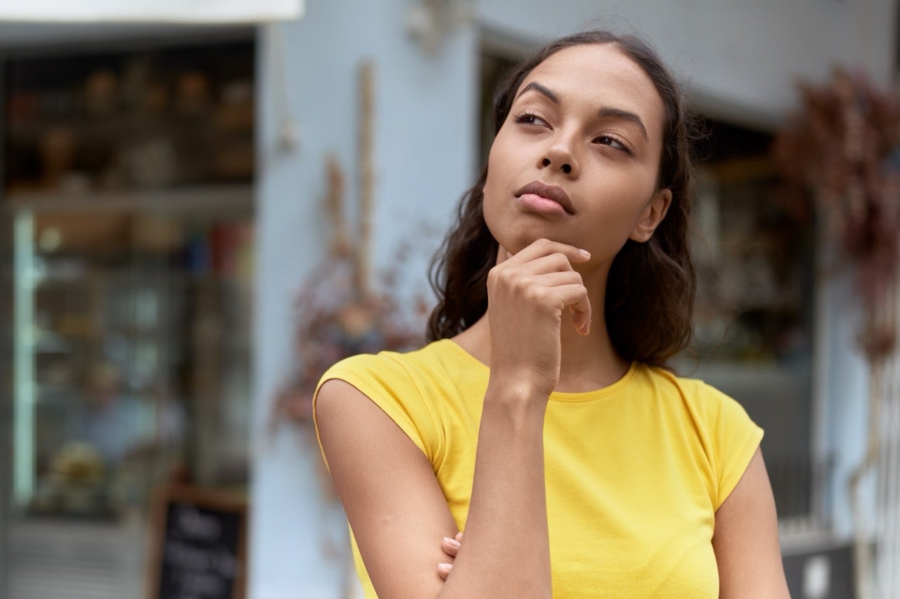 A young woman wearing a yellow shirt holds her hand to her chin as she looks into the distance. 