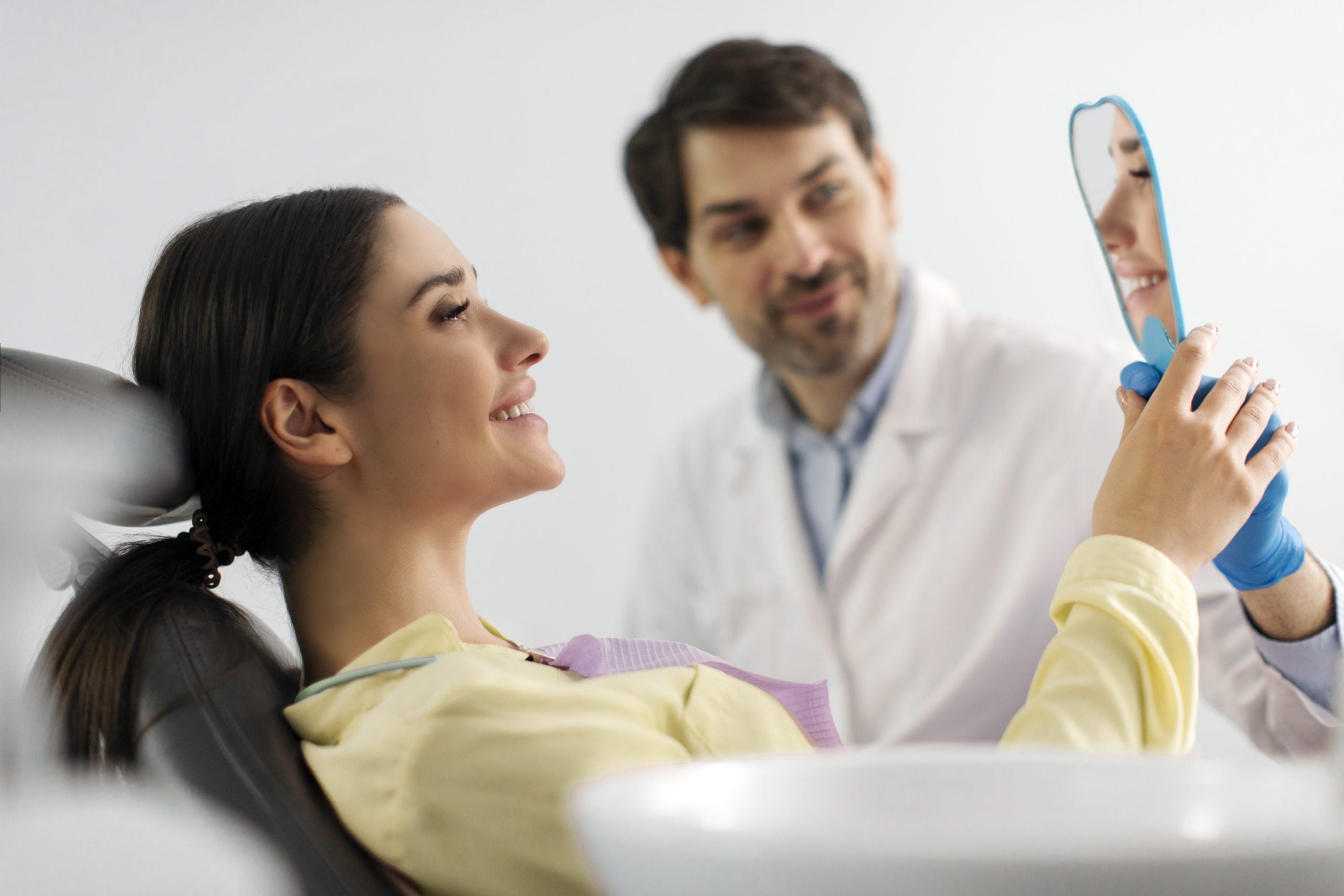 Woman reclining in dental chair smiles as orthodontist holds up mirror for her to admire her straight teeth.